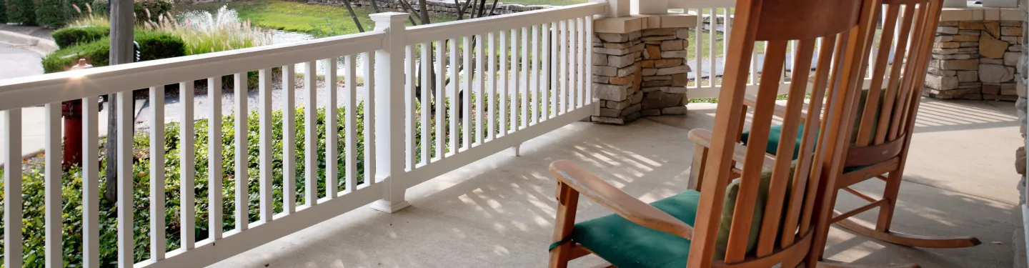 Two wooden rocking chairs with green cushions sit on a sunny, covered porch with white railings and stone columns, overlooking a landscaped yard with bushes, grass, and a sidewalk.