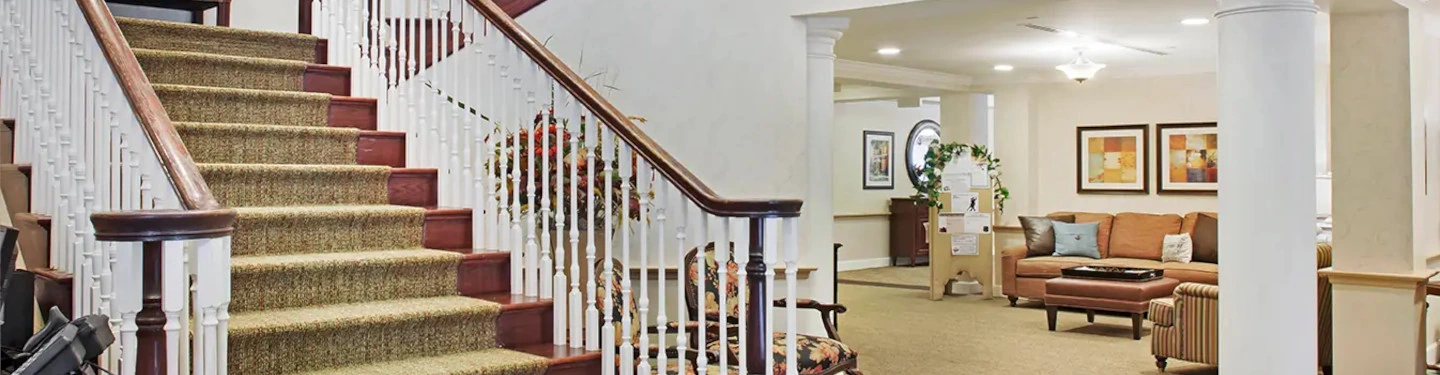 A spacious, well-lit lobby features a carpeted staircase with white railings, a cozy seating area with a tan sofa, armchairs, framed artwork on the walls, and potted plants for decoration.
