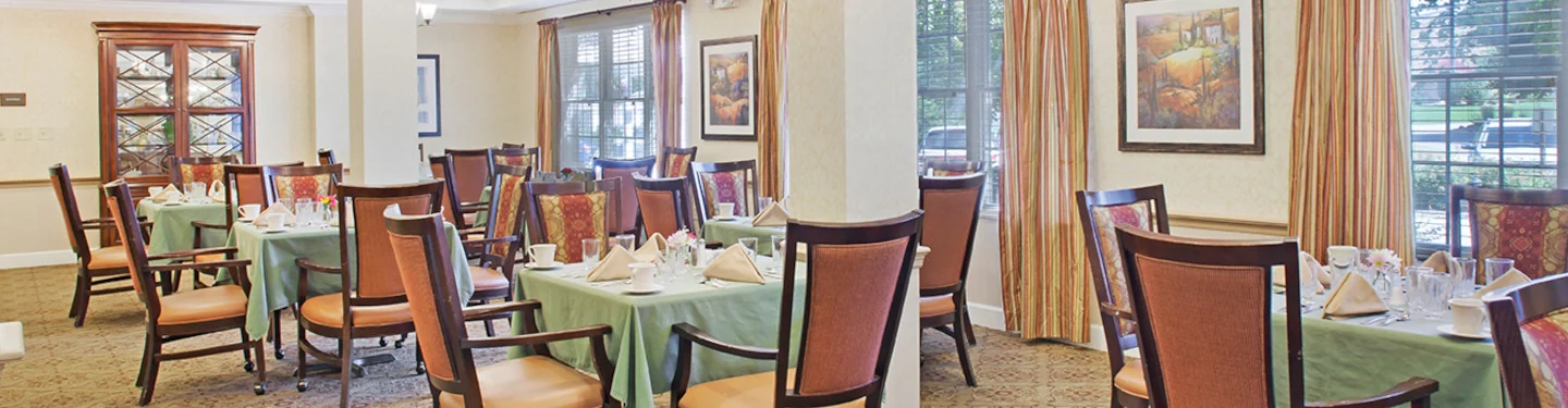 A bright, elegant dining room with several tables covered in green cloths, each set with napkins and glasses. Wooden chairs surround the tables, and large windows with curtains let in natural light. Paintings hang on the walls.