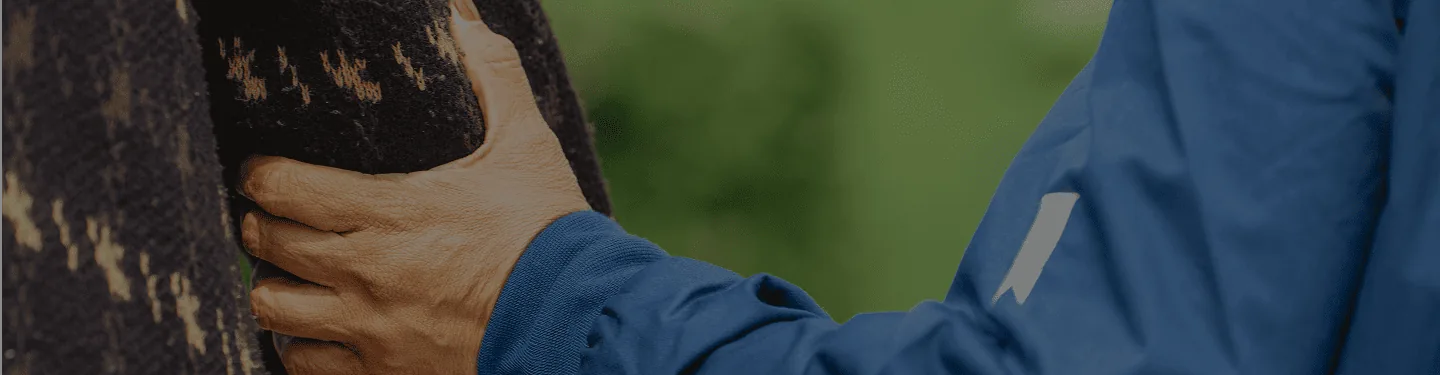 A close-up of a person wearing a blue jacket gently placing their hand on another person’s back, who is wearing a dark-patterned sweater, with a blurred green background.