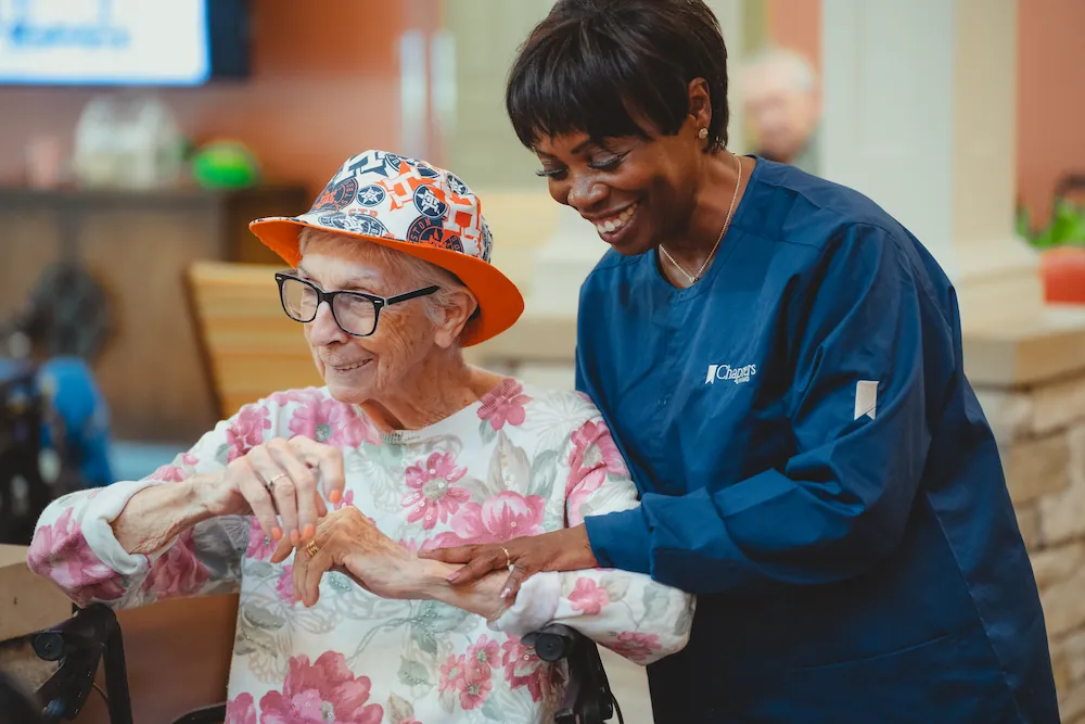 A smiling nurse in blue scrubs gently holds the hand of an elderly woman wearing glasses, a floral shirt, and a colorful hat. They are sharing a warm moment indoors.