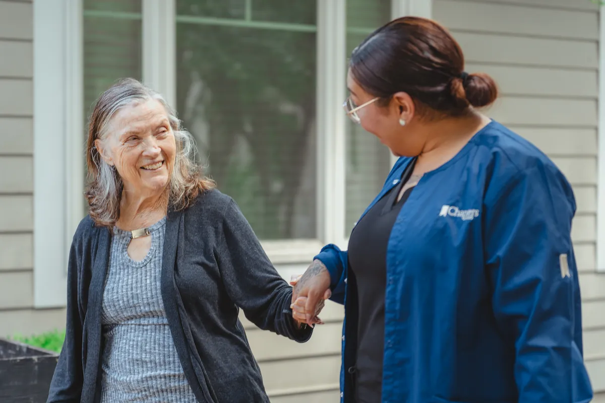 An older woman smiles while holding hands with a caregiver in a blue uniform, standing outdoors in front of a building with large windows.